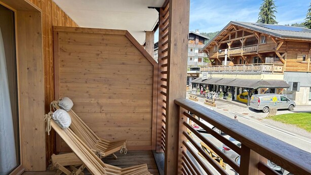 Un balcone in legno con due poltrone e vista su una cittadina con edifici e un'auto.