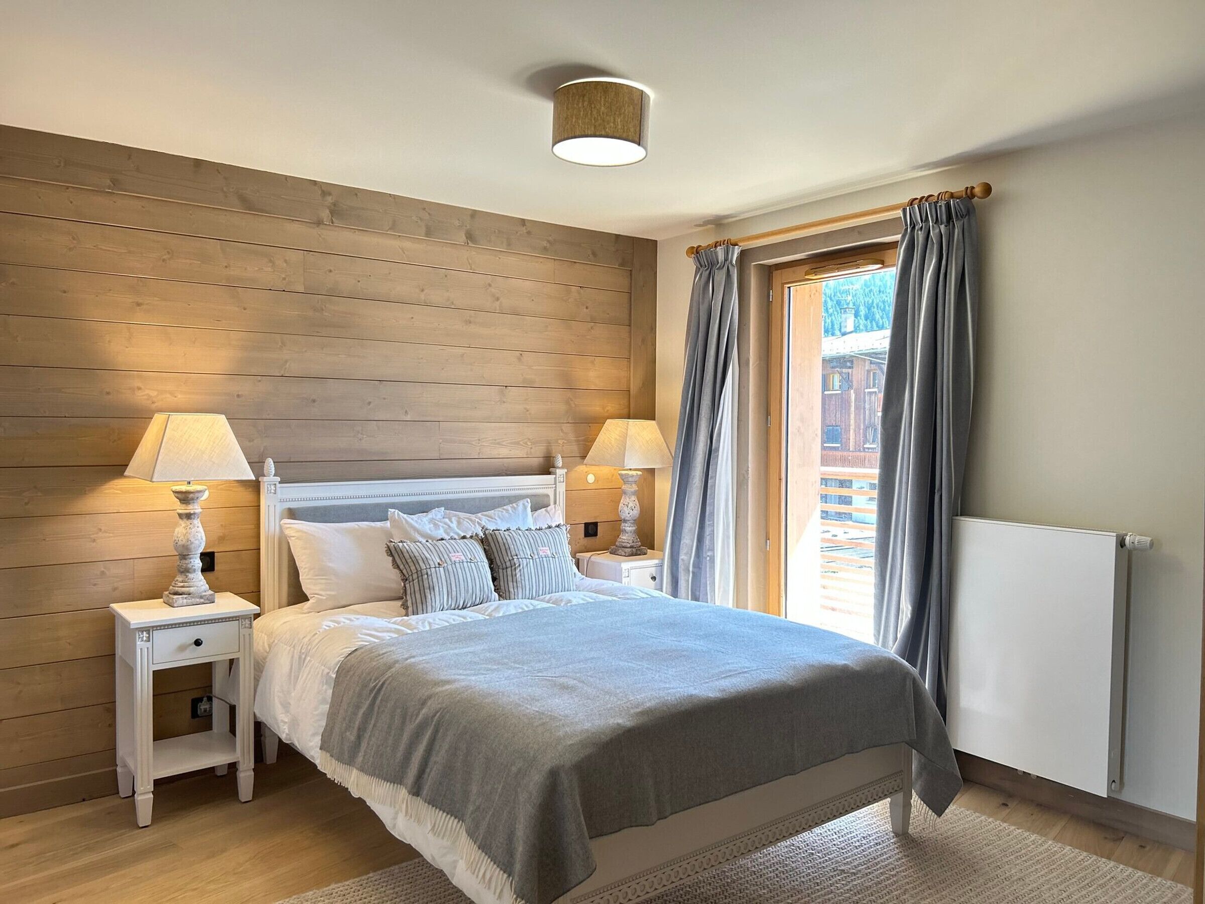 A bedroom with a wooden headboard, two bedside tables with lamps, a bed with a grey blanket, and a window with curtains.