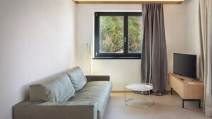 Une pièce moderne et minimaliste, avec un canapé, une petite table ronde, un téléviseur posé sur un meuble en bois et une fenêtre avec des rideaux.