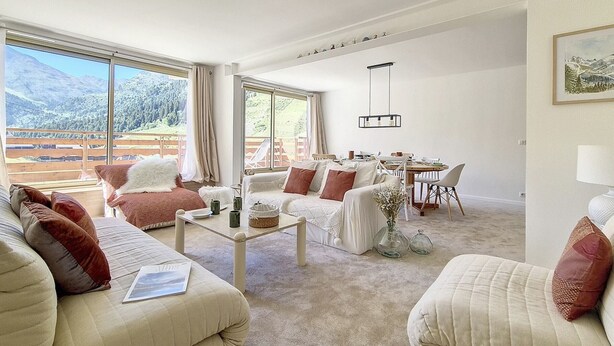 Ein geräumiges Wohnzimmer mit großem Fenster und Bergblick, einem Essbereich mit Tisch und Stühlen sowie einer gemütlichen Sitzecke mit Sofas und Kissen.