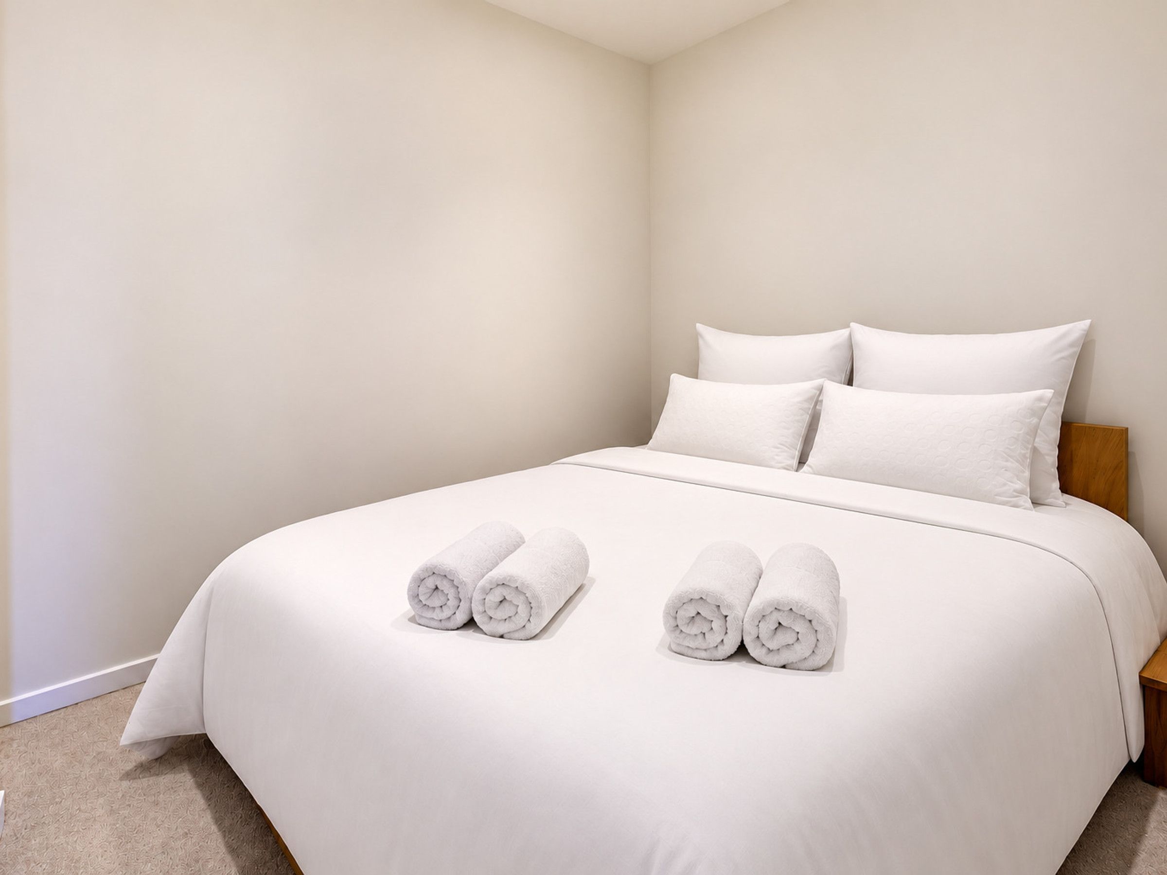 A neatly made bed with white linens and pillows, a wooden headboard, and a carpeted floor.