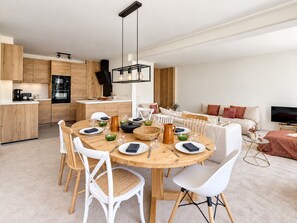 Een moderne keuken met houten kastjes, een eethoek met een ronde tafel en witte stoelen, en een woonkamer met een bank en een televisie.