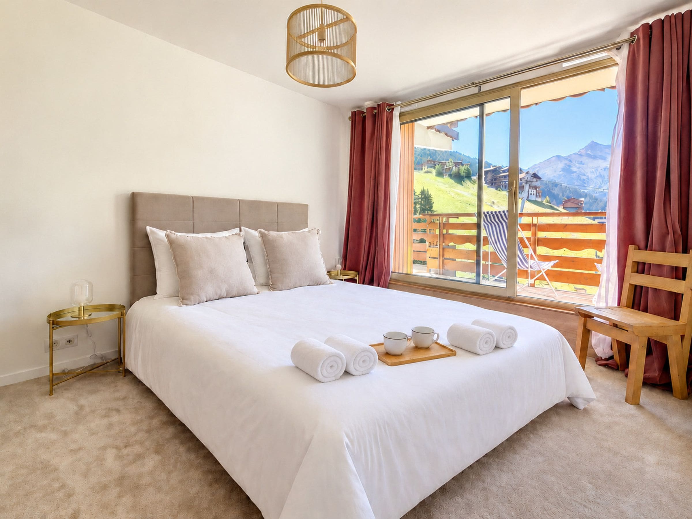 A hotel room with a large bed, a bedside table, a chair, and a window with a view of a mountainous landscape.