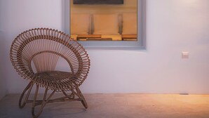 A wicker chair in front of a window with a painting, a light fixture on the wall, and a wooden ceiling beam. - Villa Cataleya (Μύκονος)