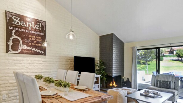 A modern living room with a fireplace, a dining area with a wooden table and white chairs, and a large window offering a view of the outdoors.