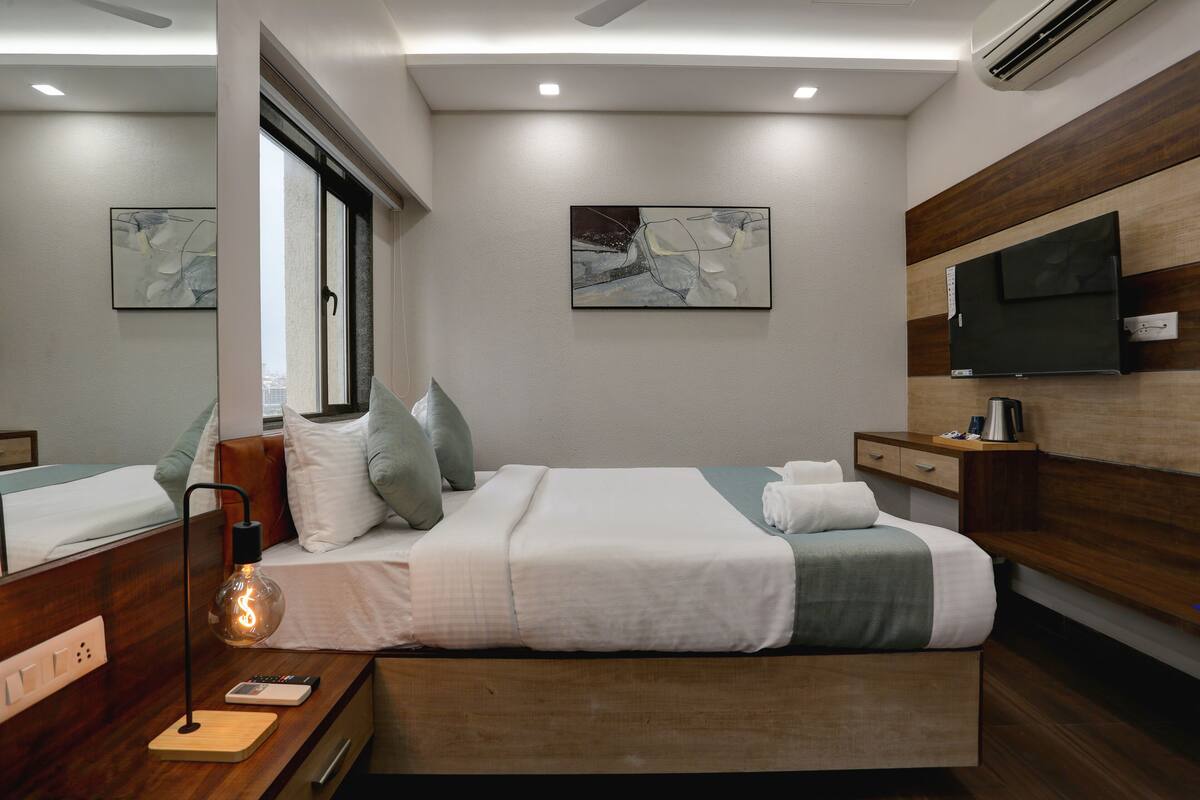 A modern hotel room with a wooden bed, a bedside table with a lamp, a flat-screen TV, and artwork on the walls.