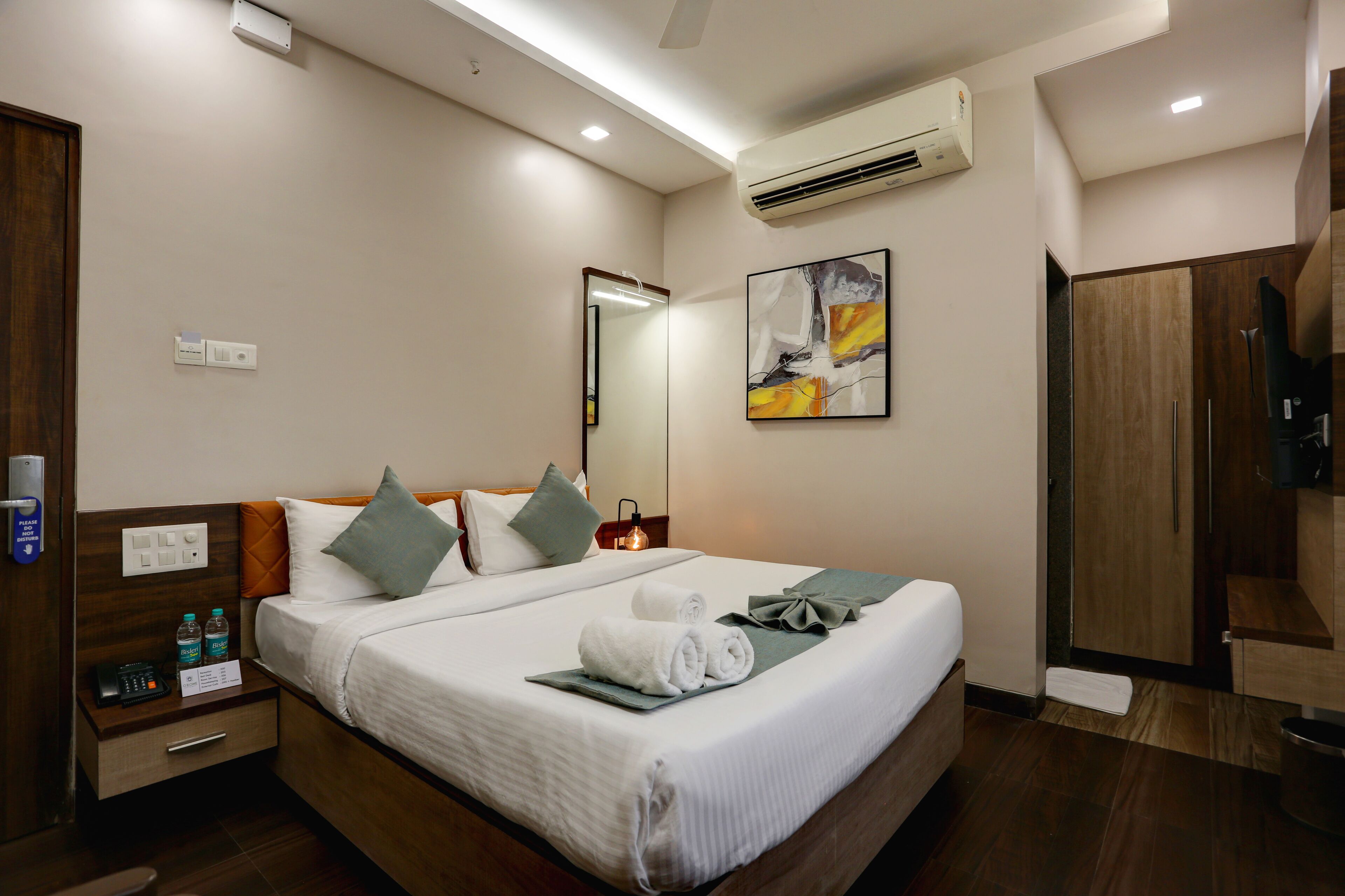 A hotel room with a neatly made bed, a bedside table with a phone and water bottles, a mirror, and a painting on the wall.