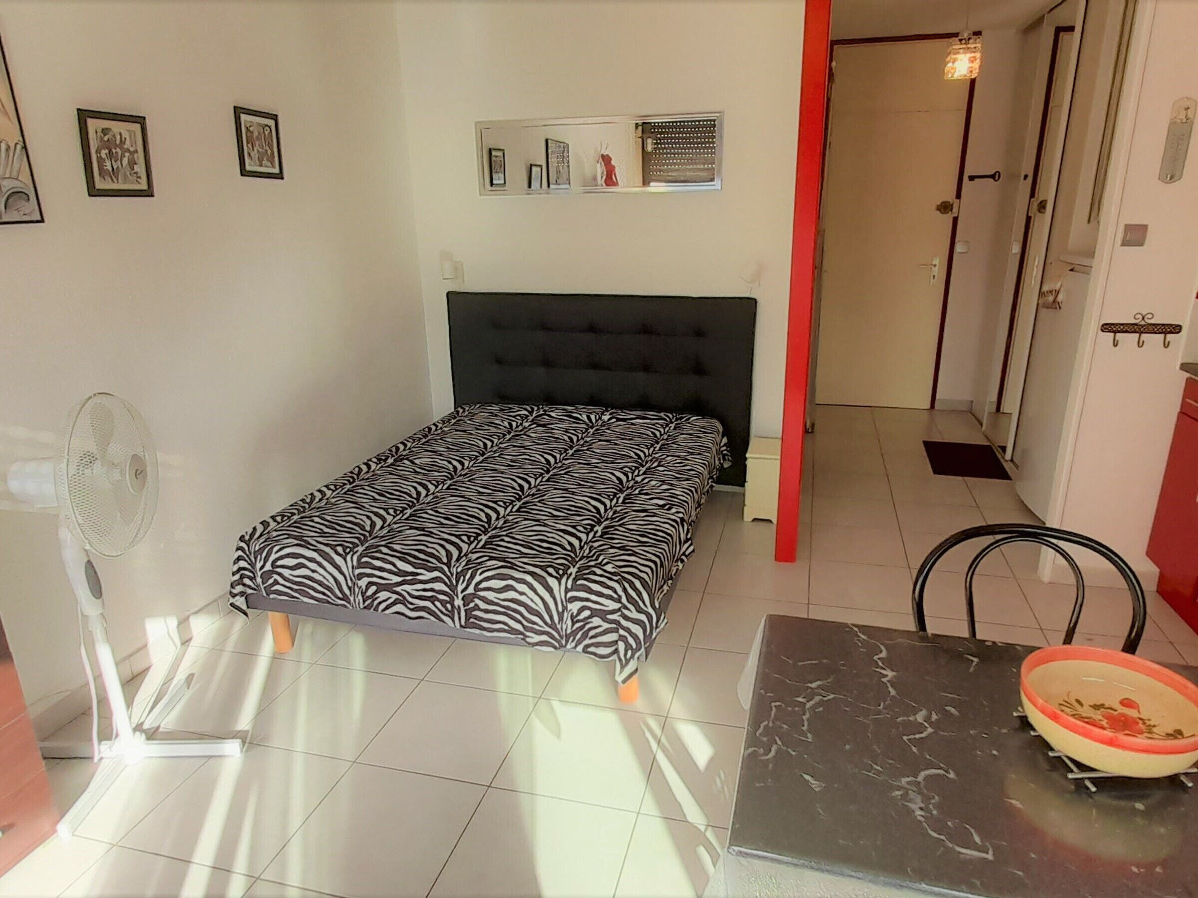 A bedroom with a zebra-patterned bed, a standing fan, a small table with a bowl, and a door leading to another room.