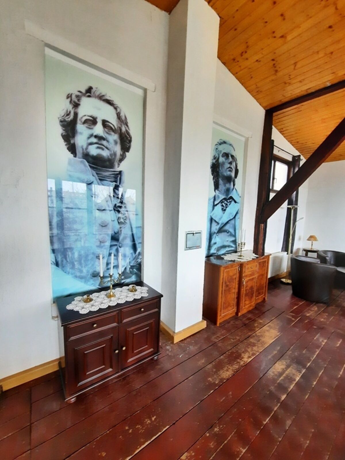 Een kamer met houten vloer, houten plafond, een kast met lades en twee aan de muur hangende schilderijen.