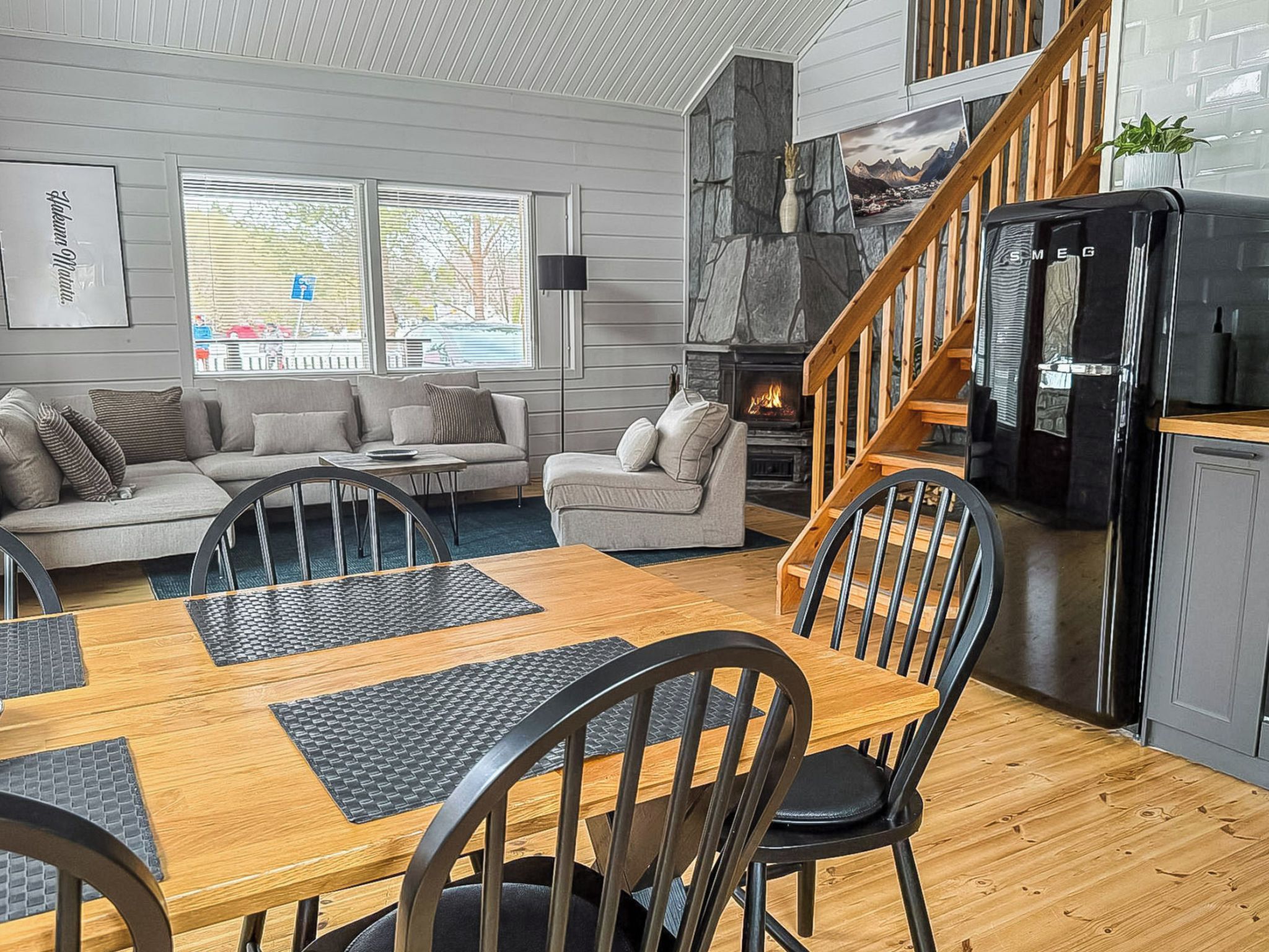 Una sala de estar moderna con chimenea, un comedor con mesa y sillas de madera, y una cocina con refrigerador negro.