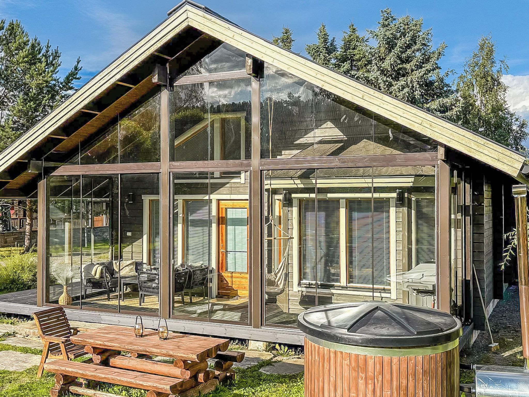 A modern glass house with a sloped roof, wooden deck, and outdoor seating.