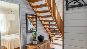 Una escalera con barandillas de madera que sube a un piso superior, una cama con cubrecama blanco, un espejo, un aparador de madera y un perchero.