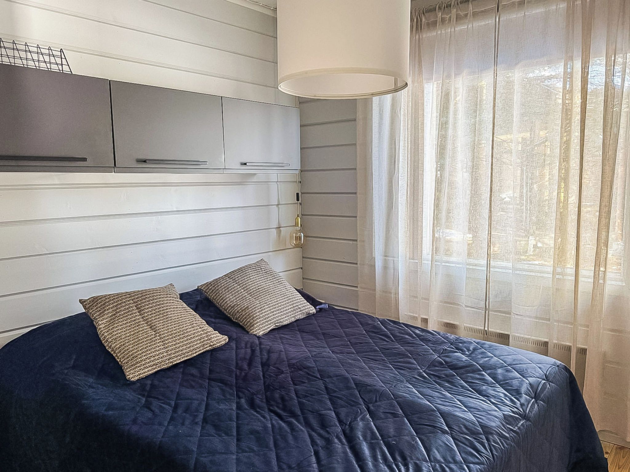 Una cama con un edredón oscuro y dos almohadas con textura, una lámpara de noche y una ventana con cortinas traslúcidas.