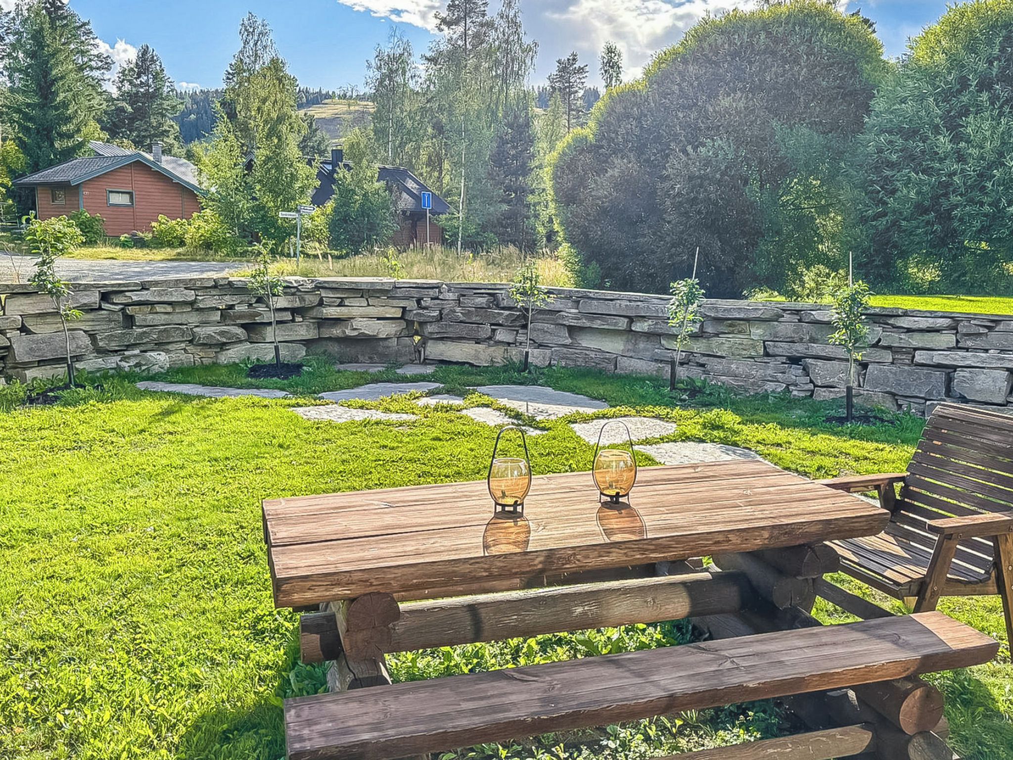 Una mesa de picnic de madera con bancos y dos faroles de cristal, ubicada en un patio trasero con césped, un muro de piedra y una casa al fondo.