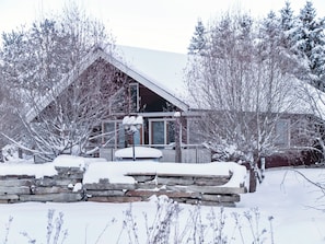 A snow-covered house with a stone wall in front, surrounded by trees.