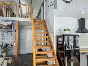 A staircase leading to a loft area, a kitchen with a modern stove, and a stone wall with a scenic painting.