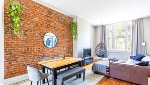 A modern living room with a brick wall, a wooden dining table, grey upholstered chairs, a brown sofa, and a hanging chair.