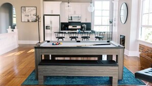 Sala de estar moderna com mesa de bilhar, cozinha ao fundo e uma grande janela que proporciona iluminação natural.