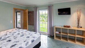Une chambre à coucher comprenant un lit, un téléviseur, une console en bois, une fenêtre avec des rideaux et une porte donnant sur l’extérieur.