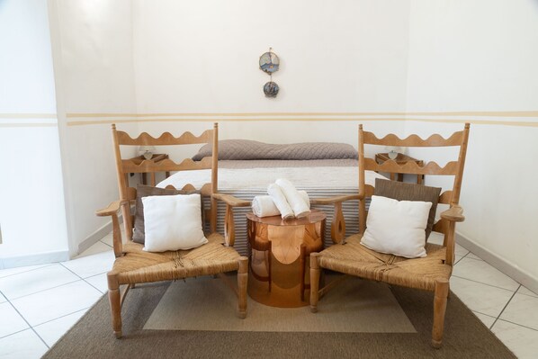 Ein Zimmer mit einem Bett, zwei Holzstühlen mit Kissen, einem kleinen runden Tisch und einer Wandleuchte.