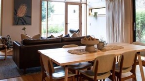 A spacious living area with a wooden ceiling, a large window, a sofa, and a dining table with chairs. - Riverstone - Perfectly positioned on the Ovens River and providing a one-of-a-kind experience (Porepunkah)