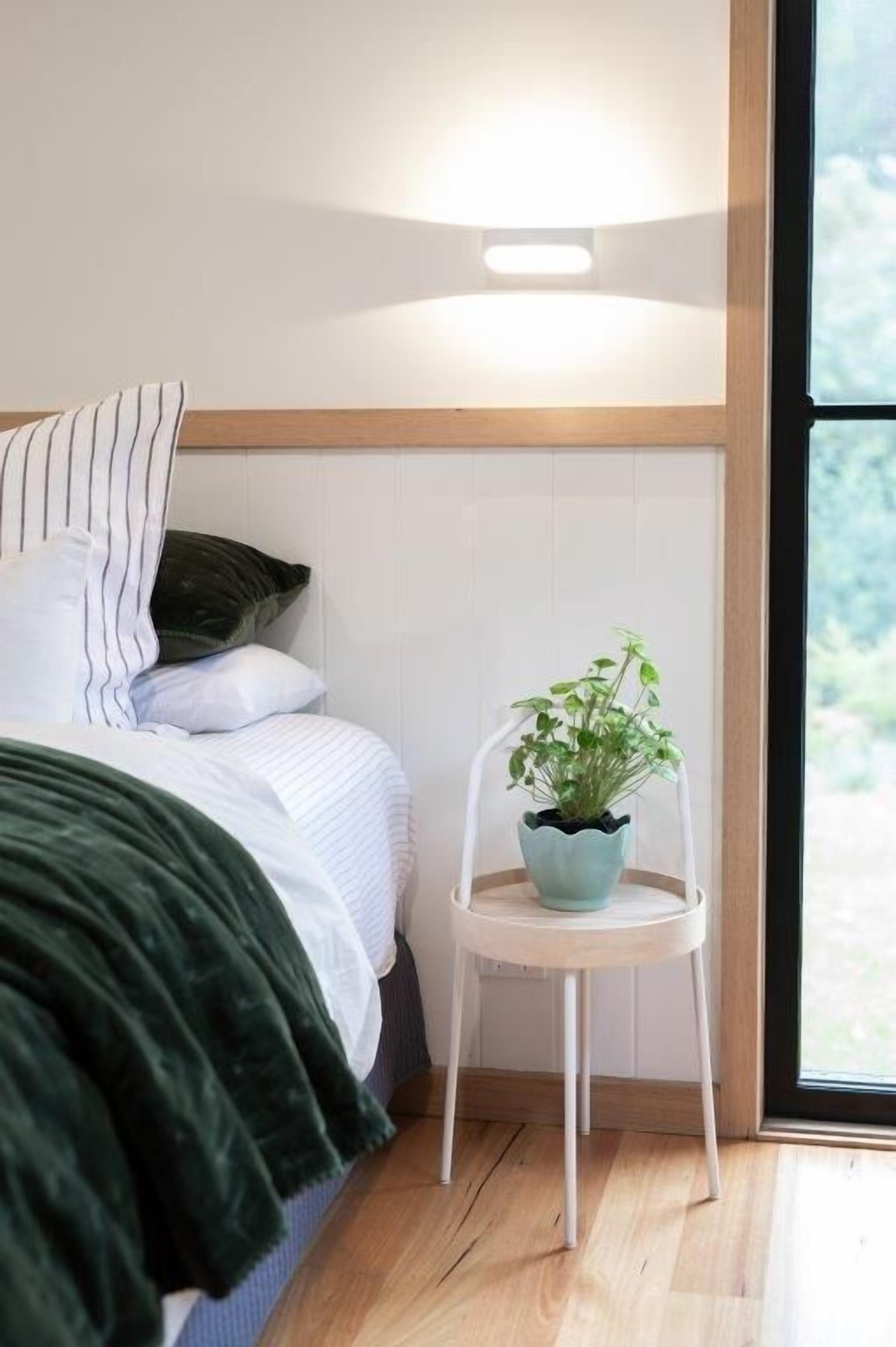 A bedroom with a bed, pillows, a green blanket, a white chair with a plant, and a window with a view.