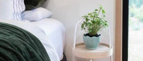 A bedroom with a bed, pillows, a green blanket, a white chair with a plant, and a window with a view.