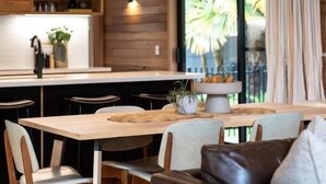 A modern kitchen with wooden cabinetry, a dining area with a wooden table and chairs, and a large window with a view of palm trees. - Riverstone - Perfectly positioned on the Ovens River and providing a one-of-a-kind experience (Porepunkah)