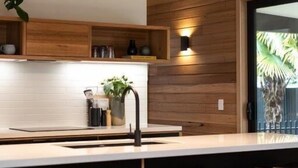 A modern kitchen with wooden paneling, a white countertop, bar stools, and a dining table. - Riverstone - Perfectly positioned on the Ovens River and providing a one-of-a-kind experience (Porepunkah)