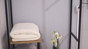 Una habitación pequeña con una cama, un estante con toallas dobladas, una mesita con una orquídea en maceta y un perchero rojo sobre una estructura metálica.