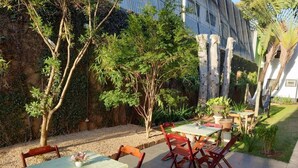 Un área de estar al aire libre con mesas y sillas, rodeada de árboles y un edificio al fondo.