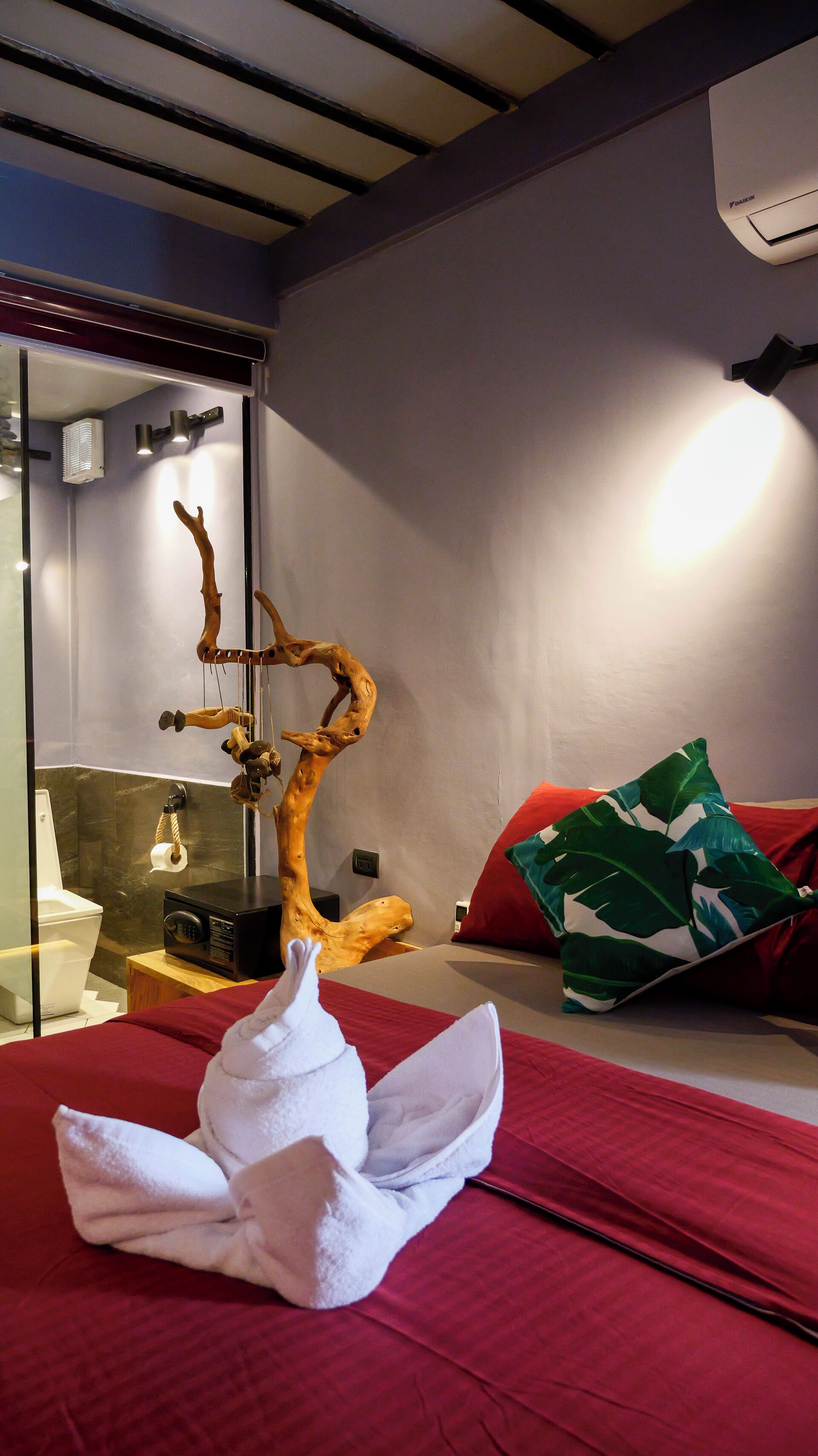 Hotelkamer met een bed, een houten sculptuur, een rood kussen, een groen kussen, een witte handdoek, een radio en een badkamer met toilet en wastafel.