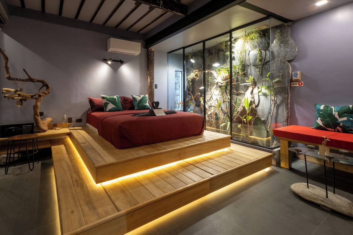 A modern bedroom with a bed, a wooden bench, a glass enclosure with plants, and a red sofa.