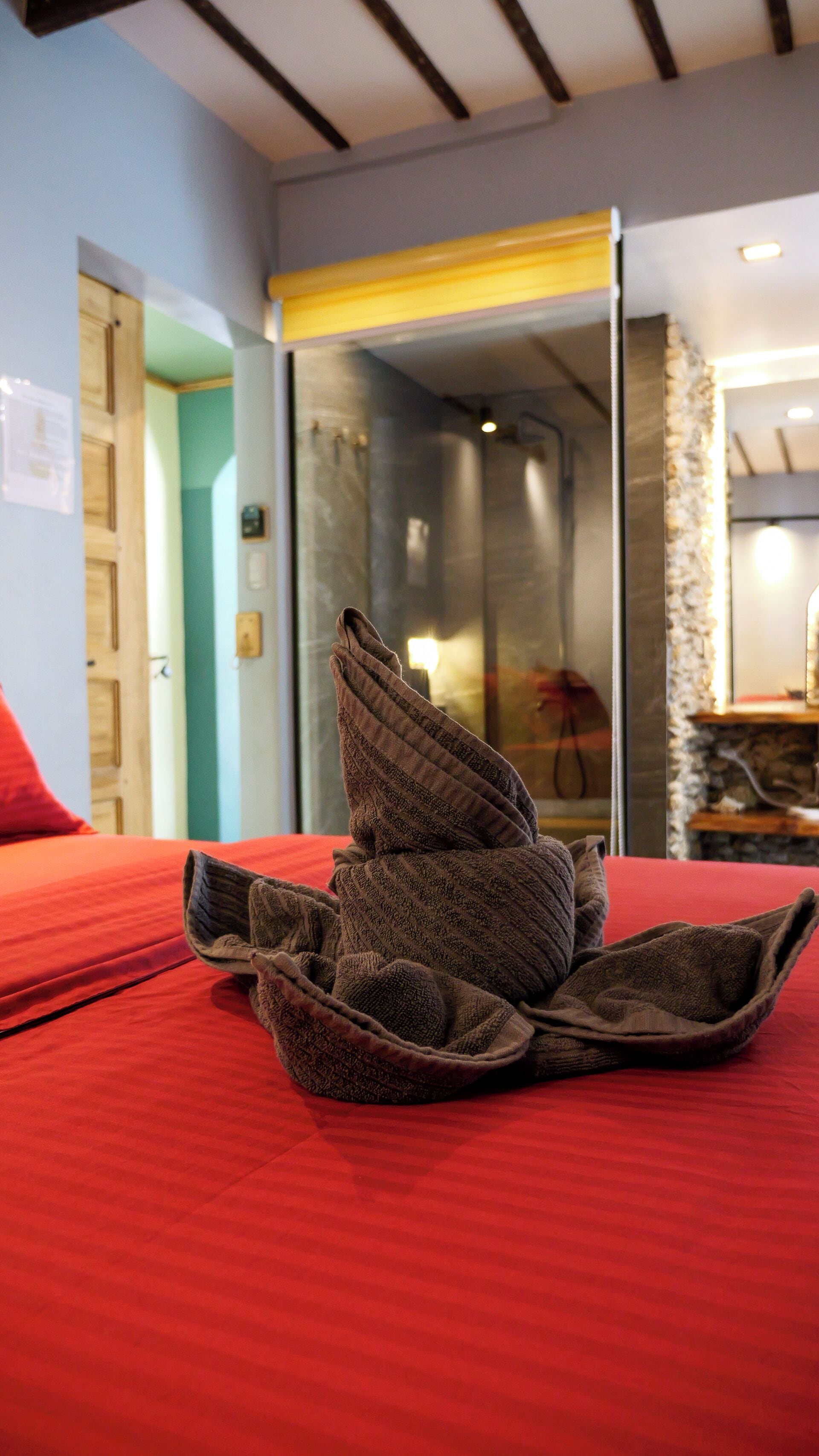 Una cama bien hecha con una cubierta roja, una toalla doblada encima y una puerta de ducha de vidrio al fondo.