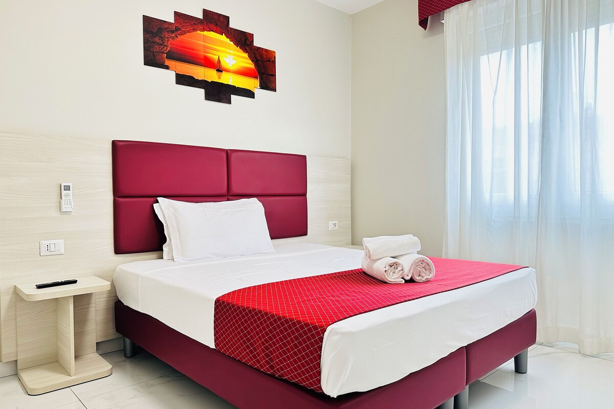 Una habitación de hotel con una cama grande, cabecera roja, mesita de noche, control remoto y una obra de arte colgada en la pared.