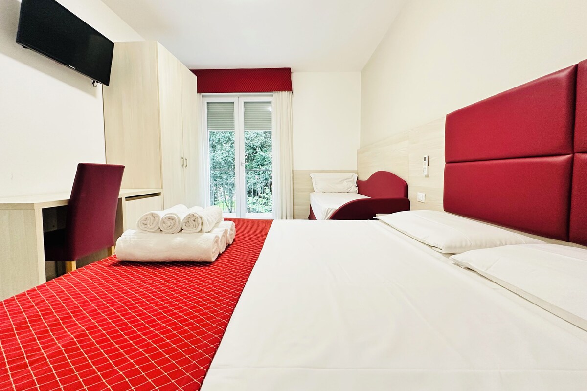 Habitación de hotel con cama, televisor montado en la pared y ventana con vistas a la vegetación.