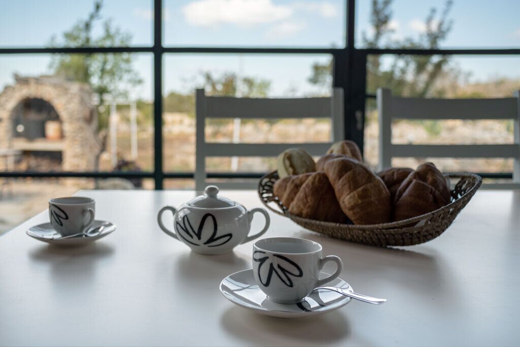 Una mesa con dos teteras y dos tazas decoradas con hojas, una cesta de cruasanes, y vistas a una construcción de piedra a través de amplias ventanas.