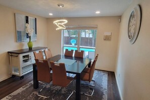 A dining area with a dark wooden table, brown chairs, a modern pendant light, a vase with white flowers, and a view of a swimming pool through a large window.