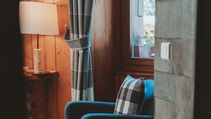 Een knus ingerichte kamer met houten vloer, een blauwe fauteuil met een geruit kussen, een lamp op een houten nachtkastje en een raam met gordijn.
