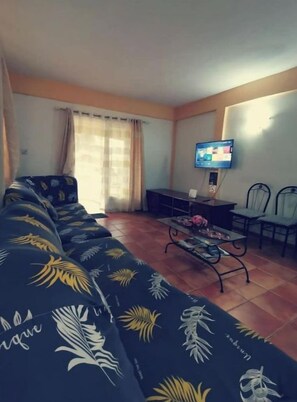 A living room with a sofa, a coffee table, a TV, and a window with curtains. - Splendide Logement de Location de 3 Chambres à Flic en Flac (Flic en Flac)