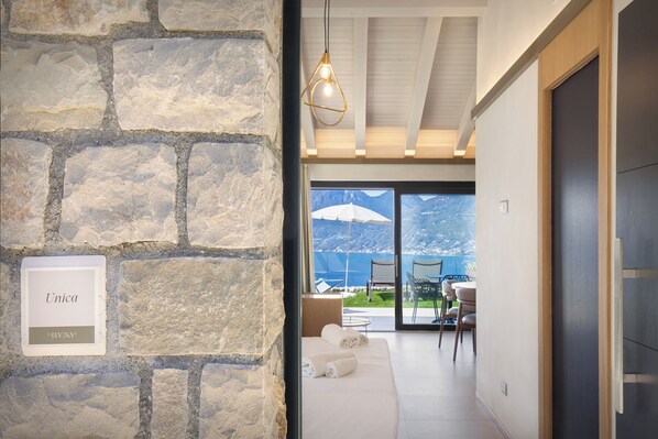 Un muro de piedra con un letrero que dice "Unica" y vistas de una habitación con una cama, sillas y una mesa, todo a través de un amplio ventanal con vistas a montañas.