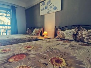 Une chambre avec deux lits, une table de chevet avec une lampe, une fenêtre avec des rideaux et un tableau floral accroché au mur.