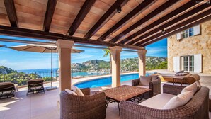 Un'area esterna coperta con mobili in vimini, una piscina e vista sul mare e sulle colline.