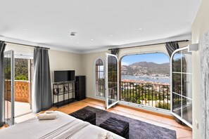 Ein Schlafzimmer mit einem großen Bett, einem Fernseher und Blick auf eine Küstenstadt durch eine Glastür.
