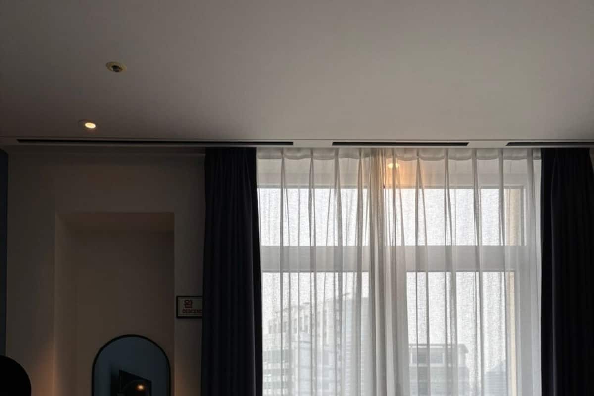 A hotel room with a bed, a small desk with a mirror, a chair, and a view of buildings through sheer curtains.