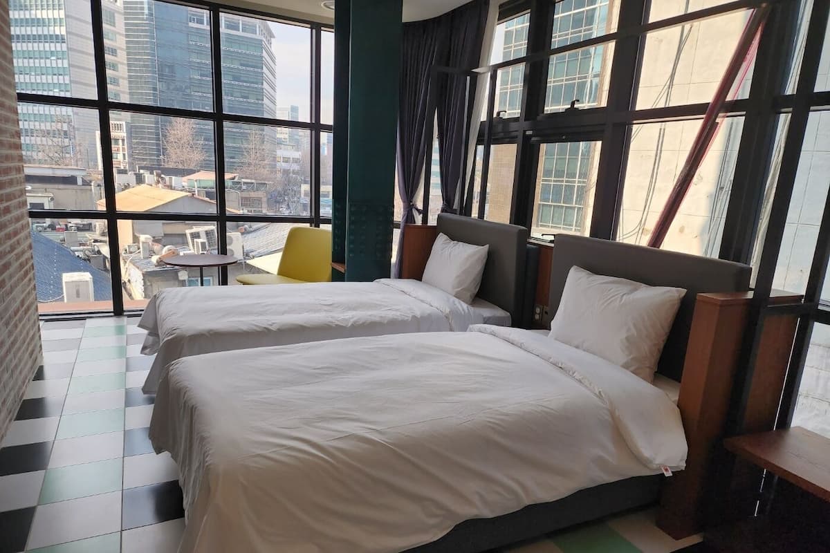 A hotel room with two beds, a view of the city through large windows, and a small table with a chair.