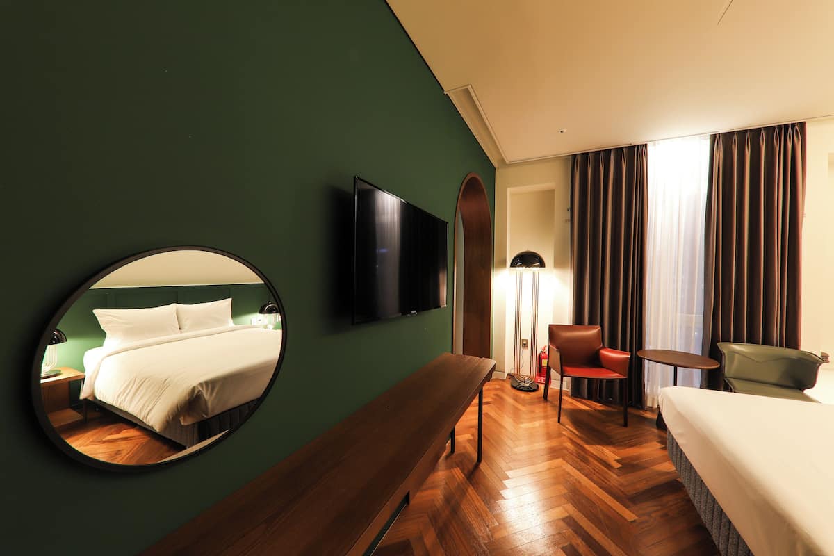 A hotel room with a bed, a round mirror, a television, a bench, a chair, and a small table.
