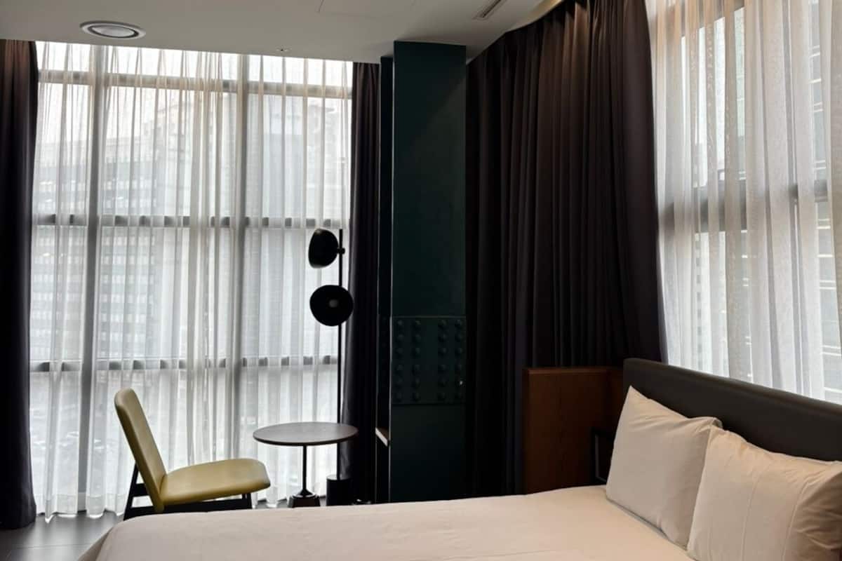 A hotel room with a large bed, a chair, a small table, and a view of the city through sheer curtains.