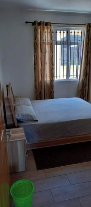 A bedroom with a bed, a window with metal bars, curtains, a green trash bin, and a small desk. - Paisible 9 Chambres à Coucher Stationnement Gratuit Pour les Vacances (Flic en Flac)