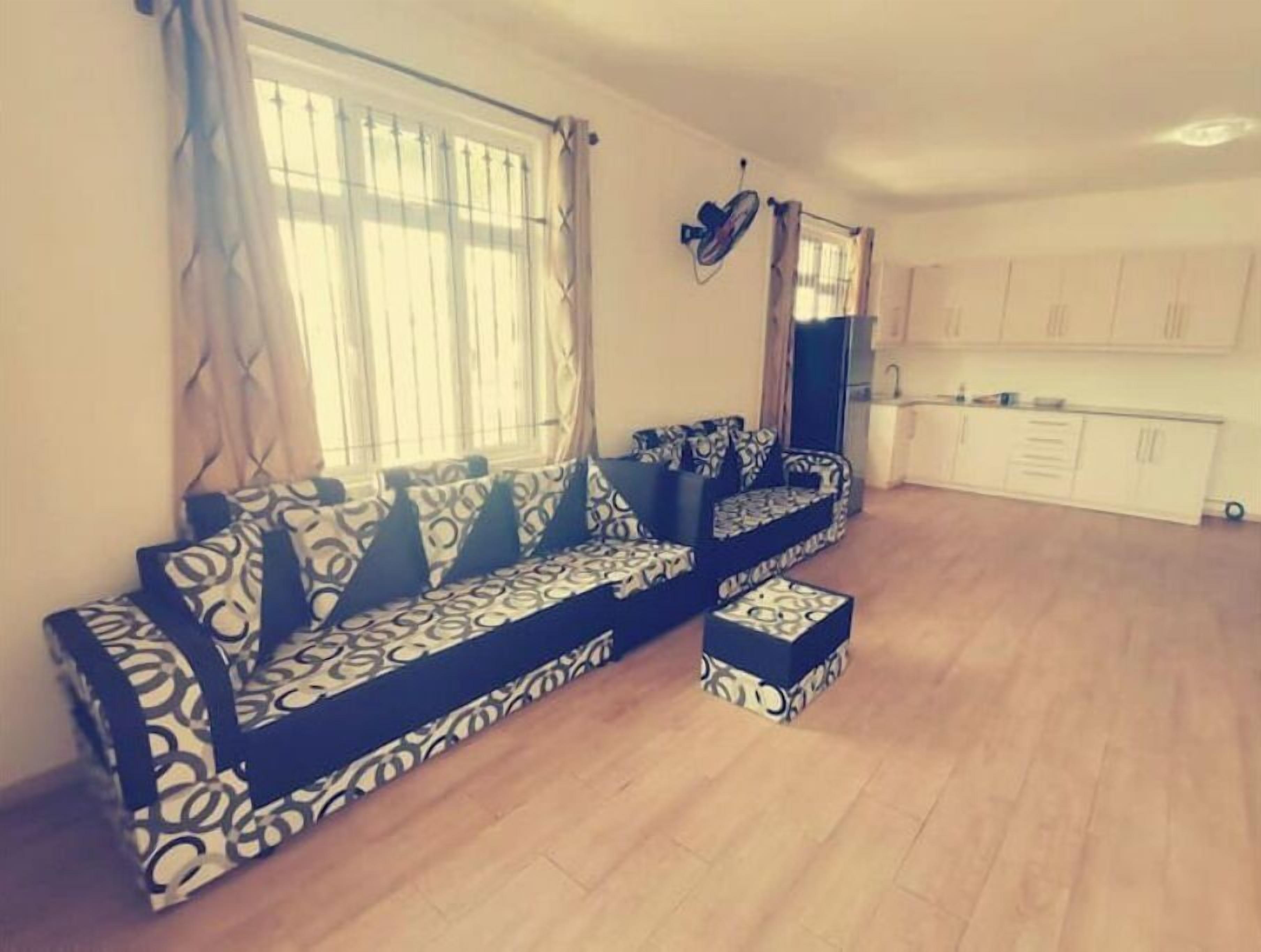 Ein Wohnzimmer mit einer Sofagarnitur und einem kleinen Hocker, eine Küchenzeile mit Schränken sowie ein Fenster mit Vorhängen.